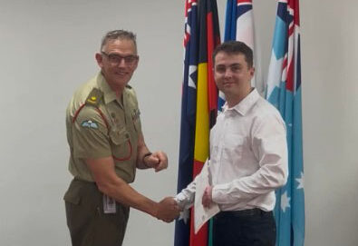Two men shake hands in a room in front of a stand of flags. One wears a tan military-style uniform and the other wears a white shirt and dark trousers while holding a sheet of paper.